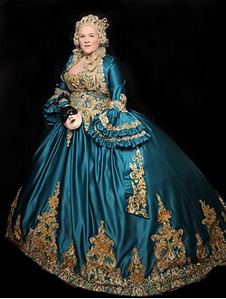 Foto zeigt Austriadine Burg in prächtiger historischen Robe wie einst Maria Theresia auf einem Gemälde. Türkises Kleid mit pompöser Gold-Bestickung, welches selbst entworfen und gefertigt wurde und weißer Perücke. Venezianische Maske in der Hand.