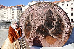Die Darstellerin von Maria Theresia, Austriadine Burg, steht vor dem 4,5 Meter hohen Maria-Theresien-Taler, welcher als Denkmal in Triest enthüllt wurde. Sie trägt ein goldenes Kleid mit Pelzüberwurf und eine weiße Rüschen Haube.